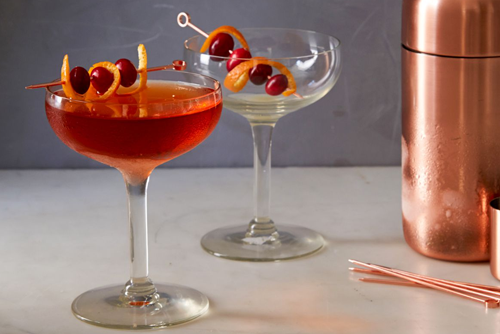 a winglass filled with a red cocktail and cherries and oranges stacked on a toothpick on the rim